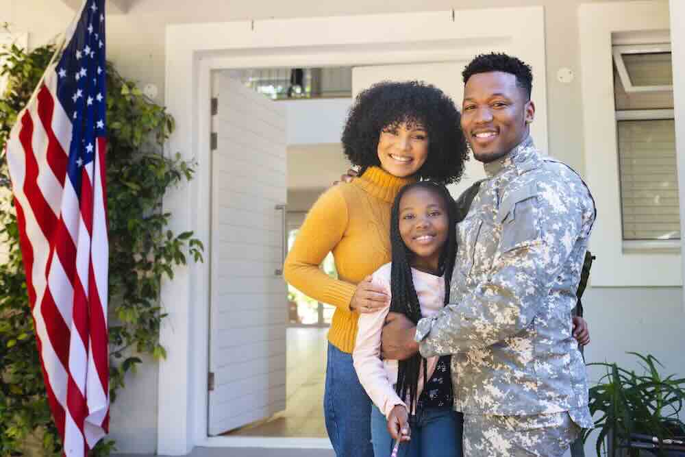 Happy family standing in front of new home with a flag to the side, representing military moves.